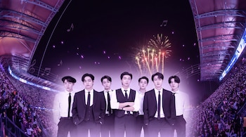 Grupo BTS posando en primer plano con trajes formales frente a un estadio lleno de fans con luces moradas, fuegos artificiales y notas musicales flotando.