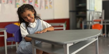 Una niña con uniforme a cuadros sonríe sentada frente a un pupitre gris en un salón de clases con sillas de colores