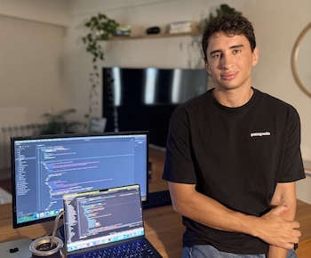 Joven sonriente con camiseta negra "Patagonia" frente a un escritorio con un portátil, monitor mostrando código y un mate