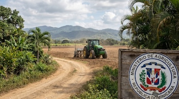 Un tractor verde y sembradora en un campo arado. Caminos de tierra en primer plano, vegetación tropical y montañas al fondo. Cartel del Ministerio de Agricultura.