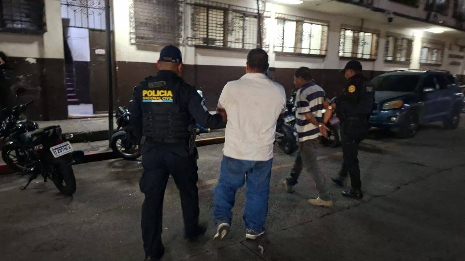 Agentes de la Policía Nacional Civil escoltan a dos hombres por una calle urbana iluminada junto a motocicletas estacionadas. (PNCdeGuatemala)
