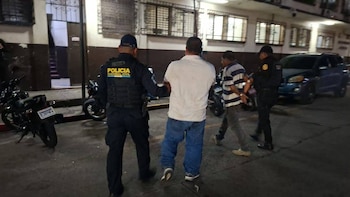 Dos agentes de la Policía Nacional Civil caminan junto a dos hombres por una calle urbana nocturna con motocicletas al fondo