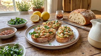 Tres tostadas con aguacate y judías blancas en un plato, junto a limones, tazones de perejil, ají y un trozo de pan sobre madera.
