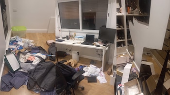 Habitación con suelo de madera lleno de ropa, cajas y papeles. Se ve un escritorio con laptop y monitor, y un espejo grande roto a la derecha