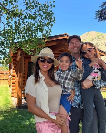 Una familia sonriente de cuatro (dos adultos, un niño pequeño y una niña) posa al aire libre frente a una cabaña de madera con fondo de montaña