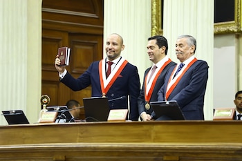 Tres hombres con traje oscuro y bandas bicolores de pie detrás de un atril, uno levanta un libro de la Constitución