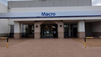 Fachada de edificio con letrero "Macro", entrada principal con puertas de vidrio, columnas, arcadas y suelo de baldosas. Dos bolardos de seguridad