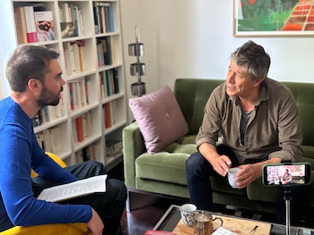 Emmanuel Carrère, de mediana edad y cabello entrecano, sentado en un sofá verde sosteniendo una taza, frente a un entrevistador joven con papeles, siendo grabado por un smartphone