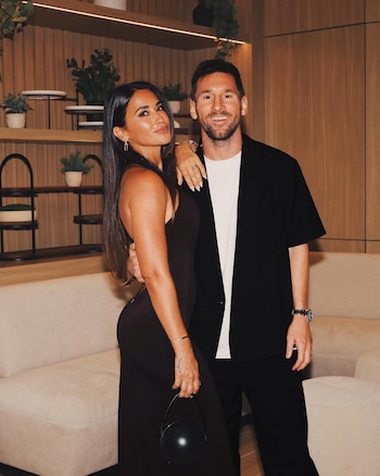 Antonela Roccuzzo, con cabello largo y un vestido oscuro, posa con un bolso esférico. Lionel Messi, con barba, sonríe a su lado