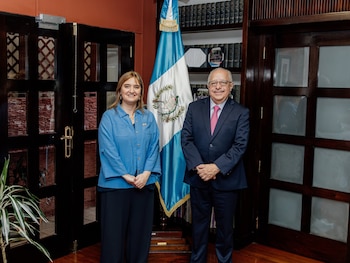Una mujer y un hombre posan de pie frente a la bandera de Guatemala. Detrás hay estanterías con libros y puertas de madera. Hay una planta en primer plano
