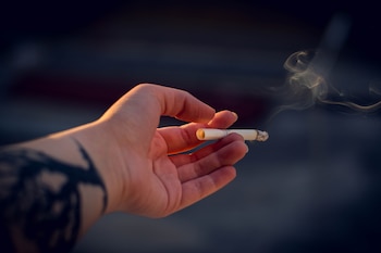 Mano con tatuaje en el antebrazo sujeta un cigarrillo encendido con filtro naranja y ceniza, del que se eleva humo. Fondo oscuro desenfocado