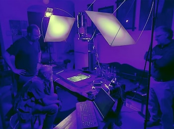 Tres hombres supervisan un proceso de digitalización de un documento antiguo en una sala con iluminación púrpura. Se ve un equipo de cámara sobre un manuscrito iluminado, un portátil y un teclado