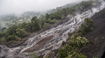 Vista aérea de una ladera volcánica con un denso río de lodo gris y marrón humeante, árboles verdes arrasados y construcciones rurales distantes entre la neblina.