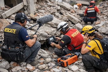 Tres personas con chalecos de rescate y cascos operan equipos de detección de sonido entre escombros. Se observa un micrófono y una pantalla con cables.