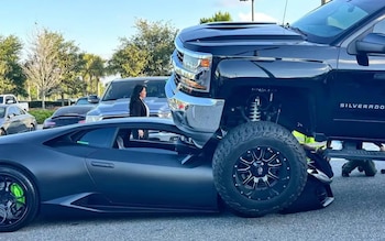 Una camioneta Chevrolet Silverado negra elevada reposa sobre un Lamborghini Huracán negro mate en un estacionamiento, mientras una mujer de cabello oscuro observa