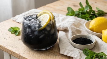 Vaso de limonada negra con hielo y rodajas de limón. Un tazón con polvo de carbón activado, limones enteros y hojas de menta en una mesa de madera.