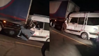 Una miniván blanca severamente dañada tras colisionar con el lateral de un camión de carga grande, cubierto con una lona azul y roja, en una carretera por la noche