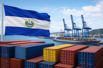 Primer plano de contenedores rojos, azules y amarillos, con la bandera de El Salvador. Al fondo, un buque de carga y grúas en el Puerto de Acajutla bajo cielo azul.