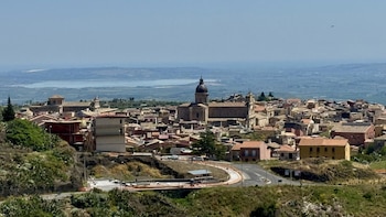 Localidad en una colina con casas de tejados rojos, una iglesia principal con cúpula oscura, carreteras, árboles y vegetación en primer plano, con un cuerpo de agua a lo lejos