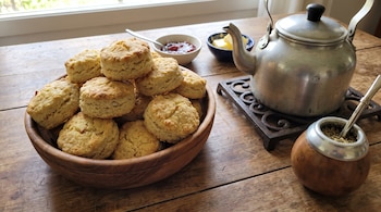 Panera de madera con scones. Junto a ella, cuencos con mermelada y manteca. Una pava metálica sobre un posafuentes y un mate con bombilla en una mesa de madera.