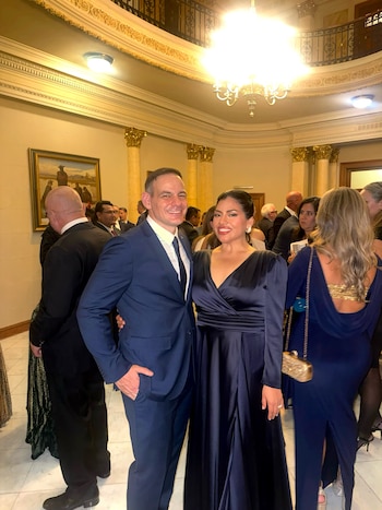 Hombre sonriente de traje azul y mujer sonriente de vestido largo azul posan. Fondo con personas, columnas doradas, lámparas y un cuadro