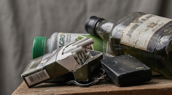 Acumulación de un paquete abierto de cigarrillos, una botella de licor, un envase de agroquímico y un aparato electrónico negro sobre madera.