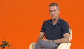Hombre de mediana edad sentado en un sillón, viste camisa polo azul oscuro y pantalones grises, con micrófono. Fondo naranja, planta y vaso de agua