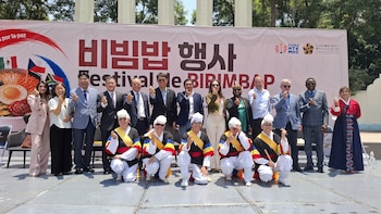 Grupo numeroso de personas sonríe y hace gestos de corazón, con algunos vestidos con trajes tradicionales coreanos, frente a un banner del Festival de Bibimbap