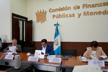 Un hombre con gafas de marco azul habla frente a un micrófono en una mesa donde hay una taza de café y una computadora portátil