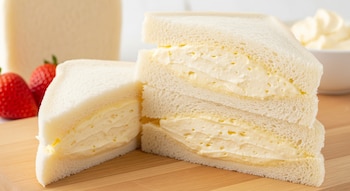 Primer plano de dos sándwiches de crema japoneses triangulares sobre una tabla de madera clara, con fresas rojas y un pan de molde al fondo.