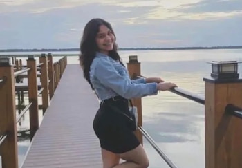 Una joven sonriente con chaqueta de mezclilla y vestido negro posa junto a la barandilla de un muelle de madera frente al agua