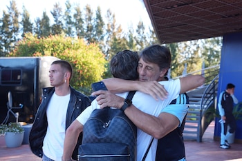 Dos hombres se abrazan afectuosamente al aire libre. Uno lleva una camiseta de la Selección Argentina. Un tercer hombre observa a la izquierda, y otro en el fondo a la derecha
