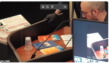 Un hombre con gafas se inclina sobre un escritorio con cuadernos y un micrófono. A la izquierda, cajas con "EVIDENCIA FORENSE". Un monitor muestra la escena del escritorio