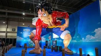 Una enorme estatua de Monkey D. Luffy, con camisa roja y pantalones azules, en una pose dinámica sobre un suelo azul. Al fondo, un mural de cielo y mar con el logo de One Piece.