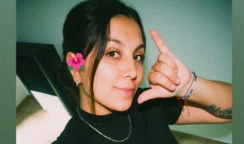 Mujer joven con cabello oscuro, flor rosa en el cabello, collar plateado, camiseta negra, pulsera y tatuaje en el brazo, haciendo una seña con la mano
