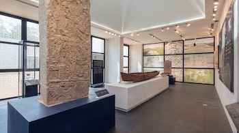 Sala de museo con gran pilar de piedra tallada en primer plano, canoa de madera sobre pedestal, y paneles en pared que muestran sitios arqueológicos y planos.