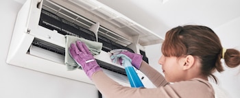 Una mujer con guantes morados limpia un equipo de aire acondicionado de pared usando un paño verde y un pulverizador