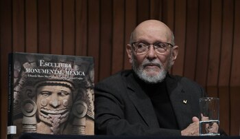 Hombre calvo con barba y gafas sentado, junto a un libro titulado Escultura Monumental Mexica con una figura tallada. Hay un vaso de agua en la mesa y una pared de madera detrás