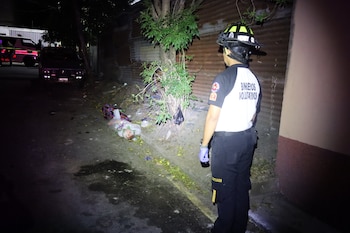 Un bombero con casco y uniforme mira un cuerpo tendido junto a un árbol en la noche. Detrás, hay un vehículo de emergencia y un coche oscuro
