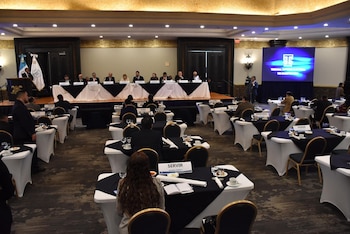 Salón de conferencias con escenario, mesa principal con panelistas, podio con banderas de Guatemala, pantalla con logo del Tribunal Supremo Electoral. Mesas con manteles, sillas y asistentes