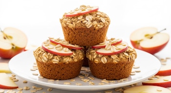 Muffins de avena y manzana apilados en un plato blanco. Están decorados con rodajas de manzana roja y avena espolvoreada. Fondo claro y luminoso.