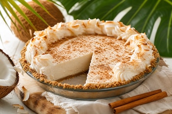 Tarta de coco sin una rebanada, con crema y canela molida, colocada en un molde de vidrio junto a cocos y varas de canela.