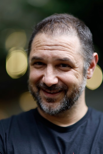 Hombre con barba y camiseta negra con un diseño rojo y gris que levanta ambas manos en un espacio con fondo desenfocado