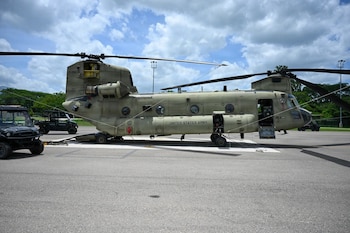 Un helicóptero militar de color verde oliva con rotores dobles y la inscripción "UNITED STATES ARMY" está estacionado en una pista, junto a dos vehículos todoterreno