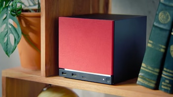 Vista frontal de una Steam Machine, un dispositivo rectangular negro con un panel rojo, sobre un estante de madera. Se aprecian puertos USB y un botón de encendido