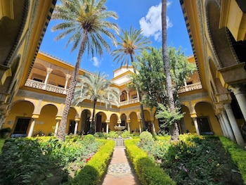 Vista del patio interior del Palacio de las Dueñas, mostrando paredes amarillas con arcos, balcones, palmeras, un jardín con flores, arbustos y una fuente