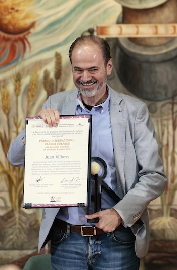 Juan Villoro sonríe vistiendo un saco gris y sostiene un diploma enmarcado junto a una escultura negra