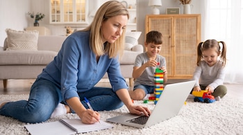 Mujer rubia sentada en el suelo con laptop y cuaderno. Un niño construye con bloques y una niña juega con un camión de juguete detrás de ella.