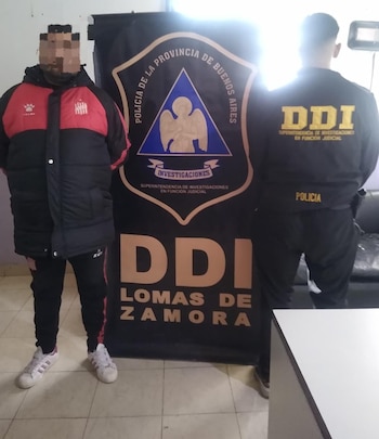Un agente de espaldas con uniforme policial negro frente a un coche Volkswagen hatchback blanco y un cartel de la DDI Lomas de Zamora