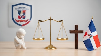 Figura de feto blanca, balanza dorada, cruz de madera y bandera dominicana sobre mesa, con logo del Tribunal Constitucional desenfocado.